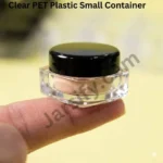 A clear, premium acrylic cosmetic jar with a glossy black cap containing a peach cream, balanced on a human index finger against a soft yellow background. Part of the 'all-in-one' 5g, 10g, and 20g empty jar collection - jarsify