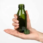 Product photo of a clear 125ml glass oil bottle with a green cap (filled as use example) standing next to an empty dark green glass bottle. Overlay text annotations show dimensions: Mouth 1 inch, Height 3.2 Inch, Base 1.5 Inch, against a light lavender background