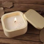 A hand holding a small, light olive green and multiple color square candle jar with rounded corners made of solid marble powder, shown with a matching square lid and a lit candle flame graphic.