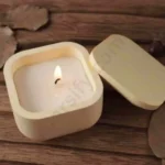 A hand holding a small, light olive green and multiple color square candle jar with rounded corners made of solid marble powder, shown with a matching square lid and a lit candle flame graphic.